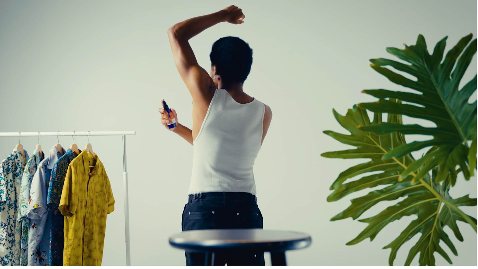 Person applying EO Citrus deodorant, seen from behind, in a room with clothes on a rack and a plant.