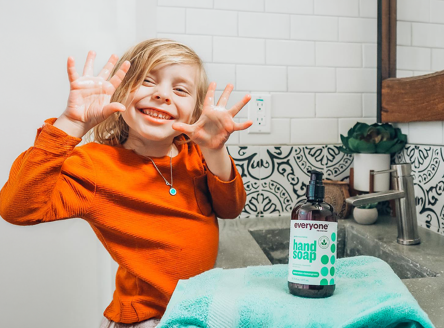 Child in an orange shirt showing off clean hands with a bottle of 'everyone' hand soap on a towel.