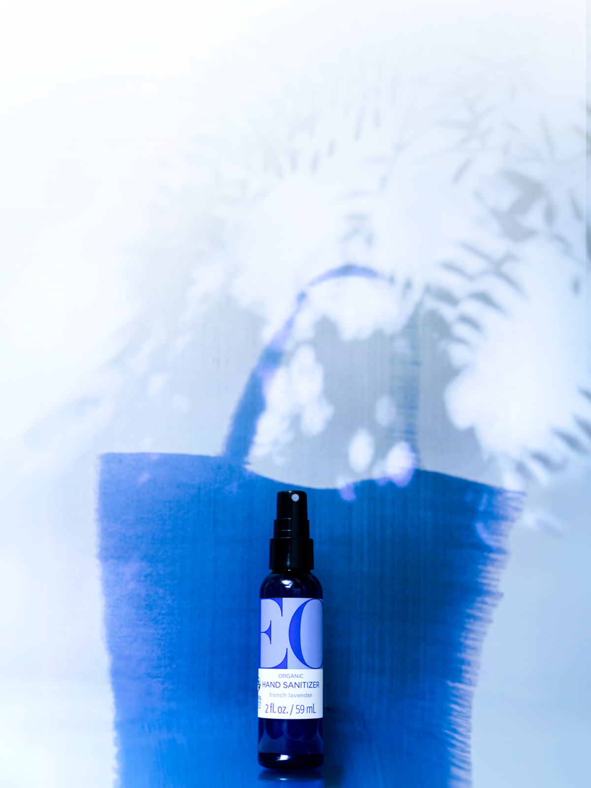 A small dark blue spray bottle labeled 'MOON GARD HYDRO SANITIZER' stands against a white background with a large blue painted brushstroke and white leaf shadows cast behind it.
